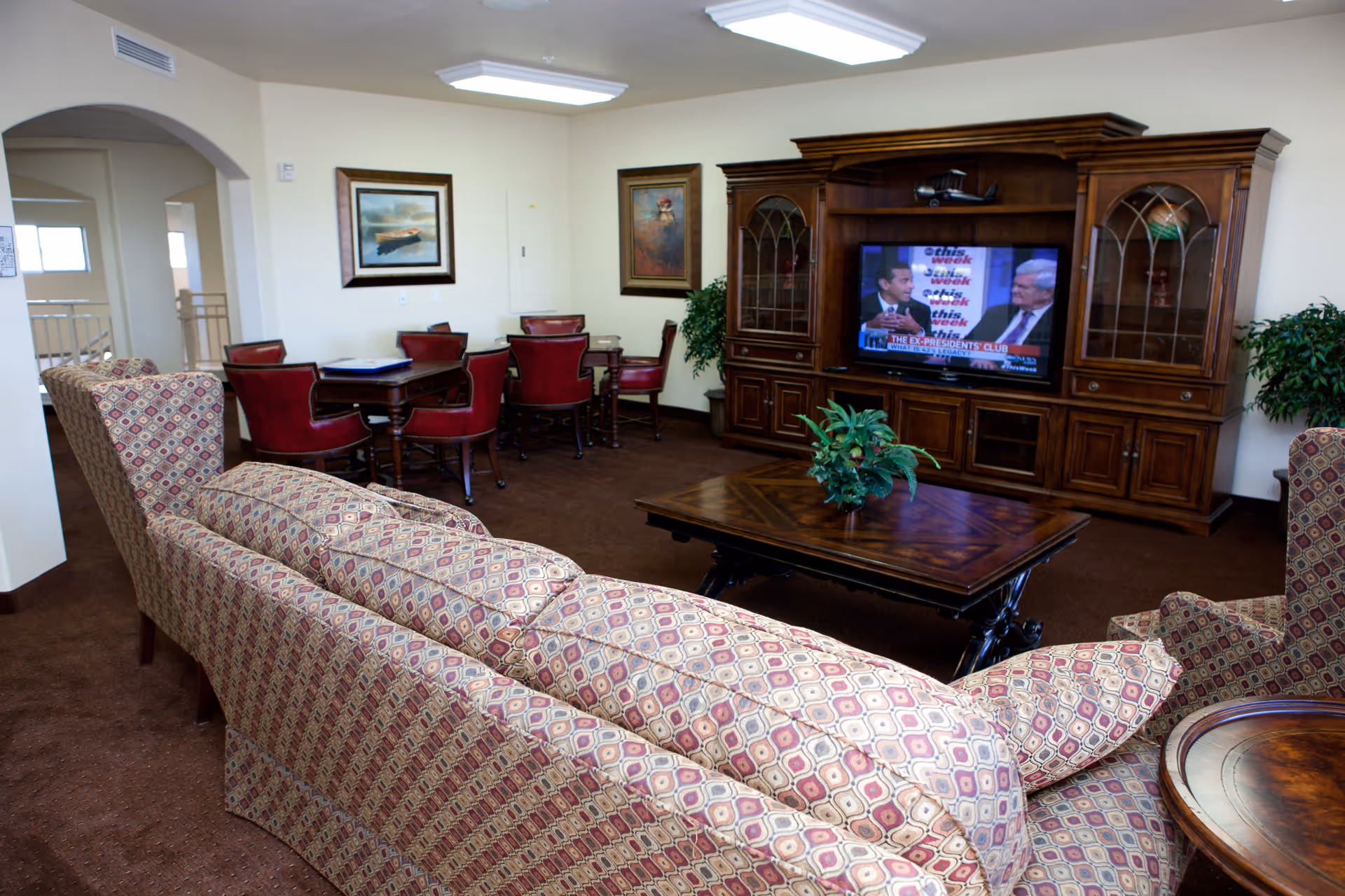 A cozy living room area with patterned upholstered sofas and armchairs facing a large wooden entertainment center with a television. The TV is showing a news program with two men in suits. There is a wooden coffee table with a small plant in the center of the room. In the background, there are two tables with red chairs and framed artwork on the walls. The room has beige walls and brown carpet.