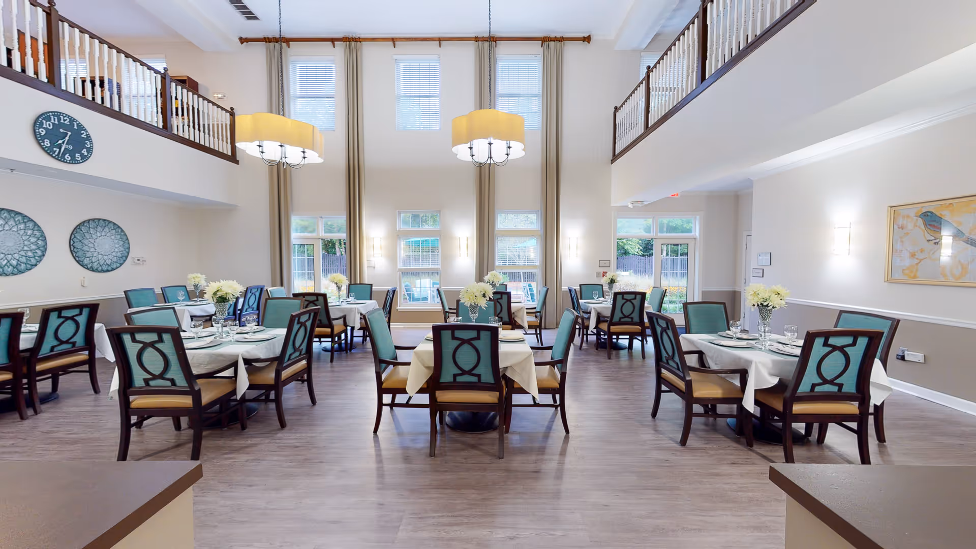 A spacious dining room with multiple round tables covered with white tablecloths, each set with plates, glasses, and floral centerpieces. The room features high ceilings with large windows letting in natural light, two large hanging light fixtures, and decorative wall art. The chairs have wooden frames with teal upholstery. There is a balcony railing visible on the upper level.