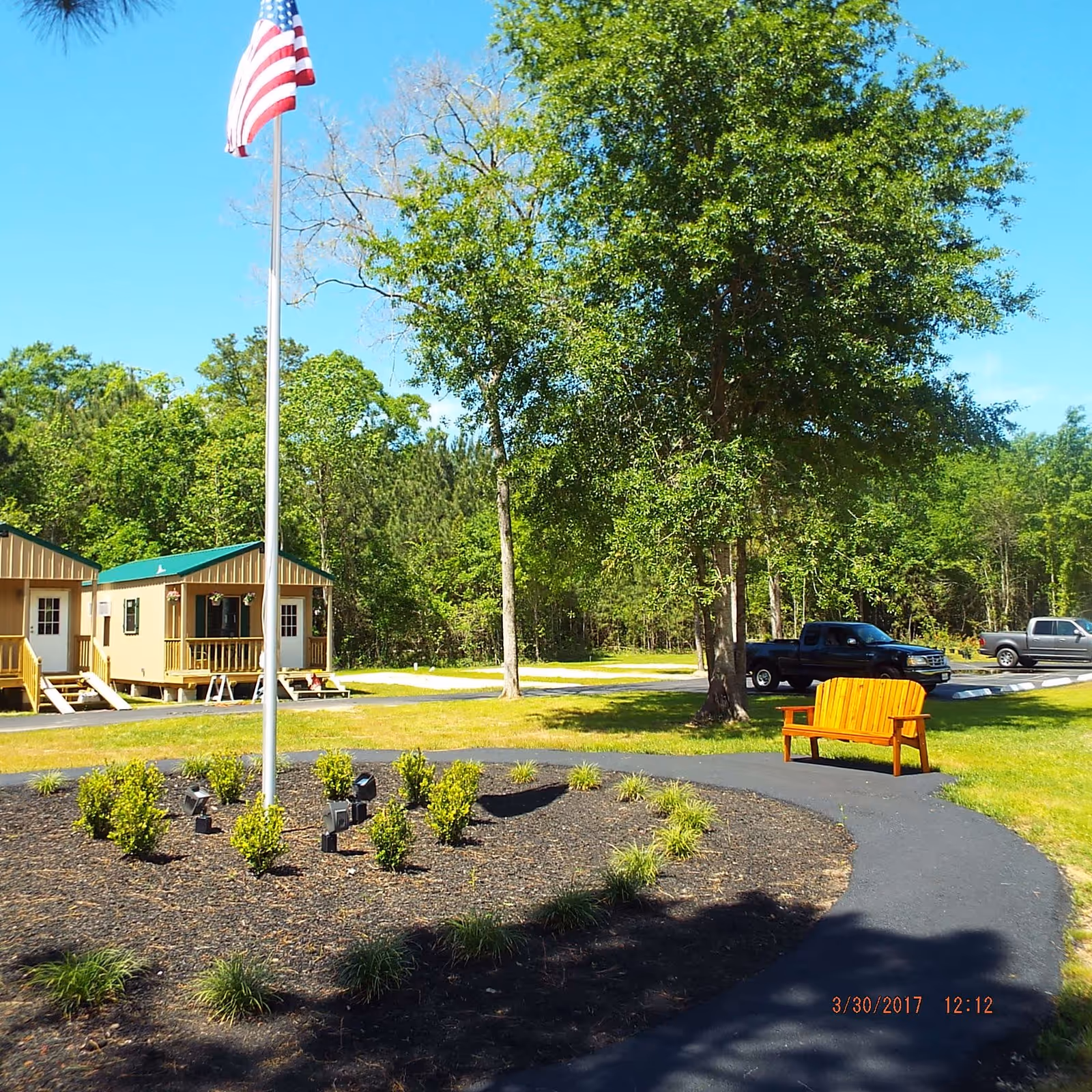 Outdoor area with a flagpole flying the American flag surrounded by small bushes and plants. There is a curved paved walkway with a wooden bench on the right side. In the background, there are two small buildings with green roofs and white doors, several trees, and two parked pickup trucks under a clear blue sky.