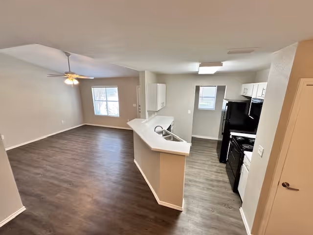 Open-plan interior showing a living area with a ceiling fan and laminate floors opening to a kitchen with a breakfast bar and black appliances.