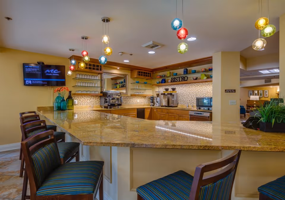 Interior view of a bistro-style kitchen area with a large granite countertop bar surrounded by wooden chairs with striped cushions. The kitchen features wooden cabinets, a coffee machine, a microwave, and shelves with colorful bowls and cups. Colorful pendant lights hang from the ceiling, and a television is mounted on the yellow wall to the left.