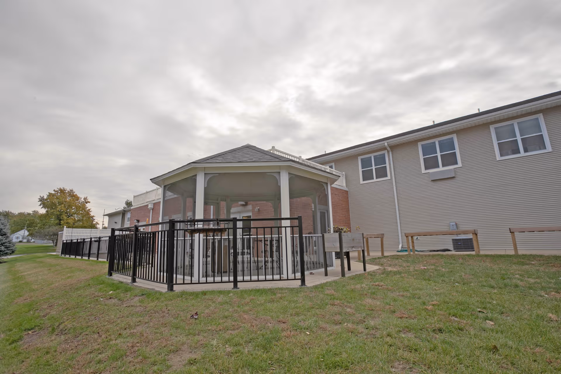 Outdoor view of a senior living facility showing a fenced gazebo with seating inside, adjacent to a two-story building with multiple windows. The sky is cloudy and the grass is green around the structure.