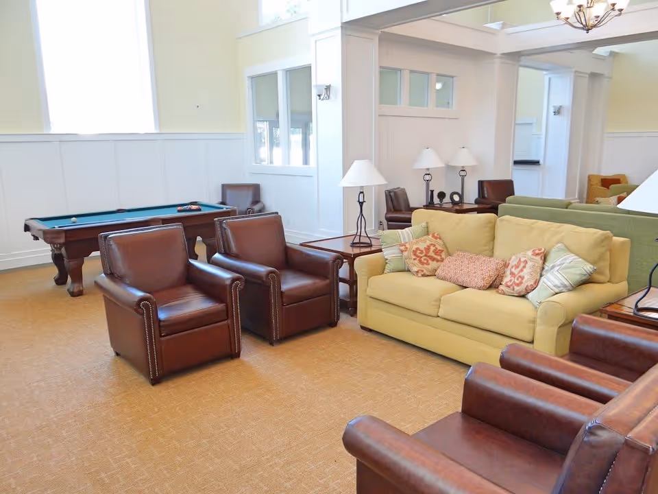A bright and spacious living room area with a yellow sofa adorned with multiple patterned cushions, surrounded by several brown leather armchairs. In the background, there is a pool table and additional seating. The room features large windows allowing natural light to fill the space, white paneled walls, and beige carpeting.