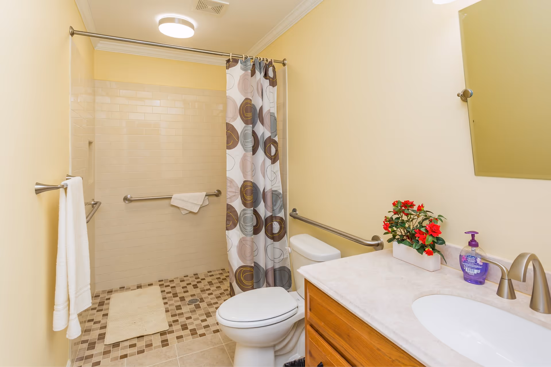 A clean bathroom with beige walls and tiled floor. It features a walk-in shower with a patterned shower curtain, a white toilet with a grab bar beside it, a wooden vanity with a white countertop, a sink, a faucet, a small potted plant with red flowers, a bottle of hand soap, a mirror, and a towel hanging on a wall-mounted towel rack.