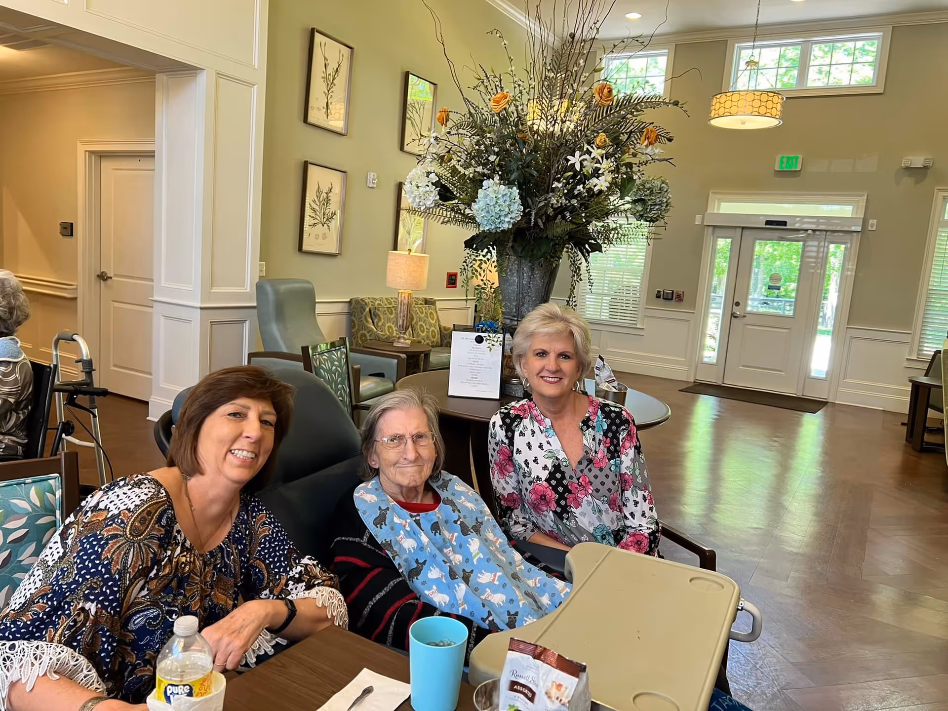 Three women sitting together at a table in a well-lit common area of a senior living facility. One elderly woman in the center is wearing a bib with cat designs, flanked by two smiling women. The room features large windows, a tall floral arrangement on a round table, framed botanical artwork on the walls, and comfortable seating.