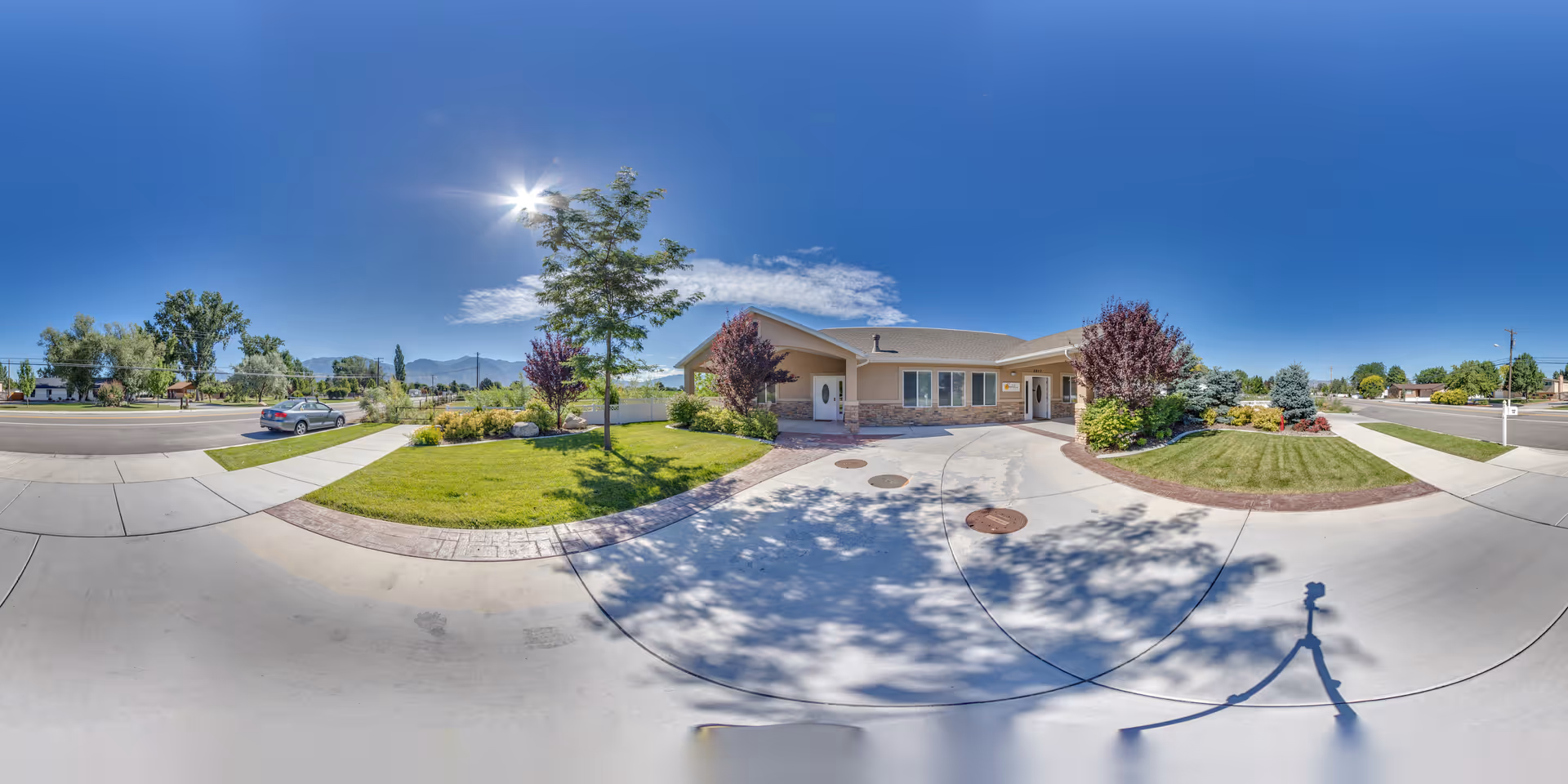 Wide panoramic view of the front exterior of a single-story residential building with a well-maintained lawn, trees, and shrubs under a clear blue sky with the sun shining brightly.