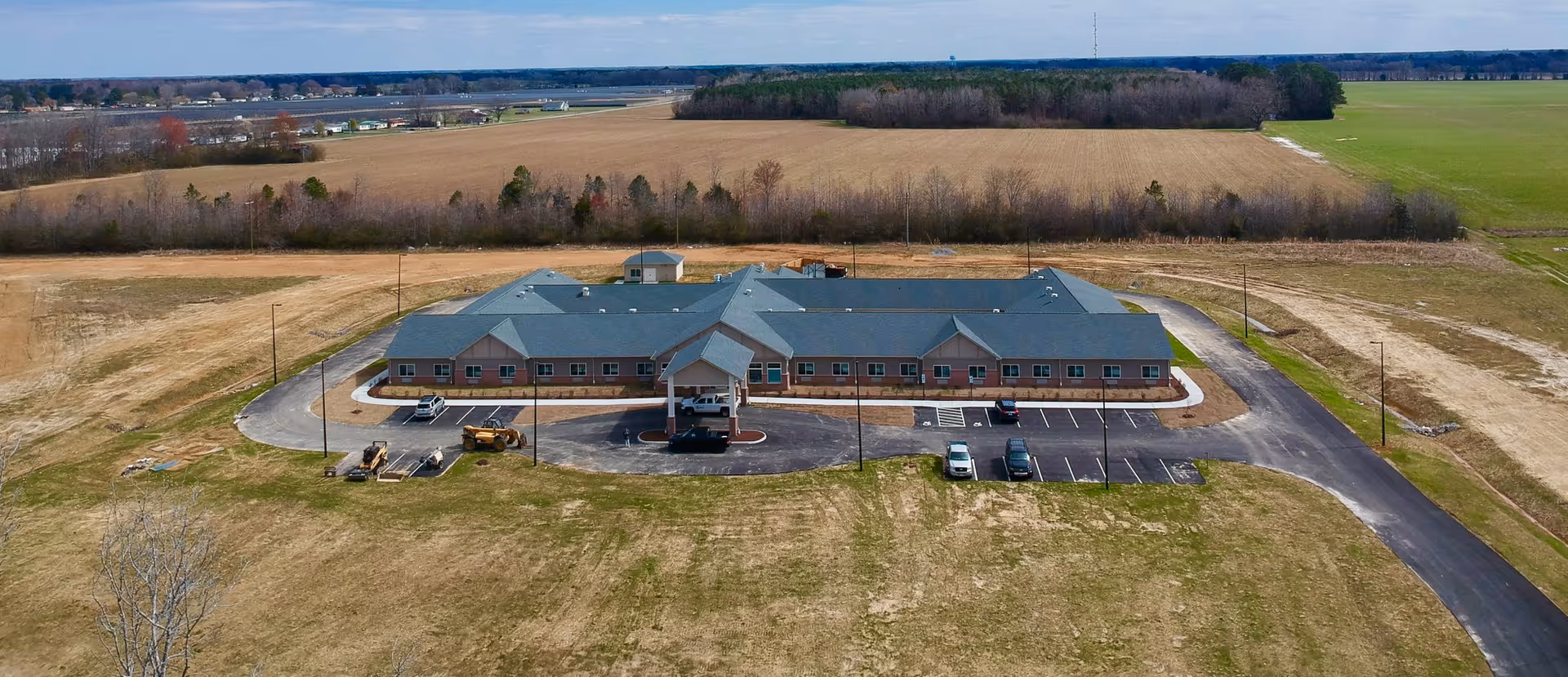Aerial view of a single-story building with a large parking area surrounded by open fields and trees in the distance under a clear sky.