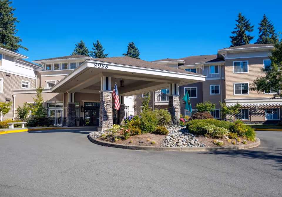 Front entrance of a multi-story senior living building with a covered porte-cochere, flags, and a landscaped roundabout.
