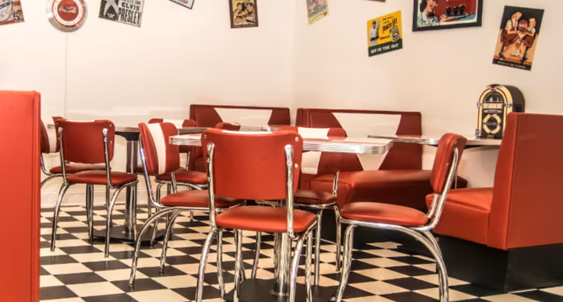 A retro-style dining area with red and white cushioned booths and red chairs with chrome legs around silver tables. The floor has a black and white checkered pattern. The walls are decorated with vintage posters and a small jukebox is placed on one of the tables.