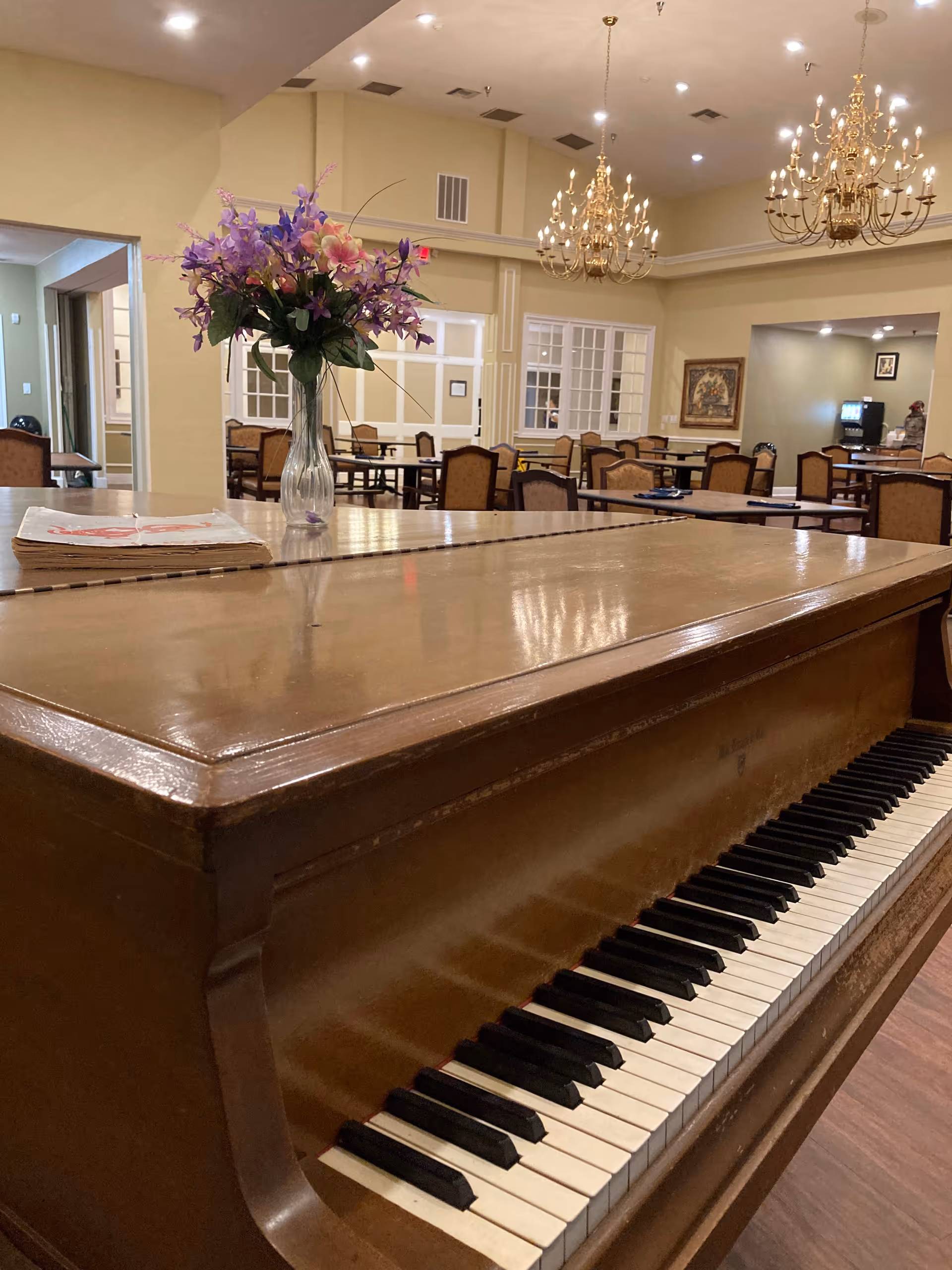 A wooden piano with a vase of purple and pink flowers on top, situated in a spacious dining area with multiple tables and chairs. The room features beige walls, large windows, framed artwork, and elegant chandeliers hanging from the ceiling.