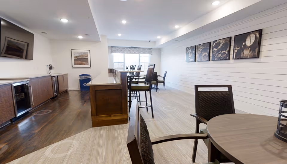 Bright senior living common room with tables and chairs, a central island/bar, wall-mounted TV and framed artwork.