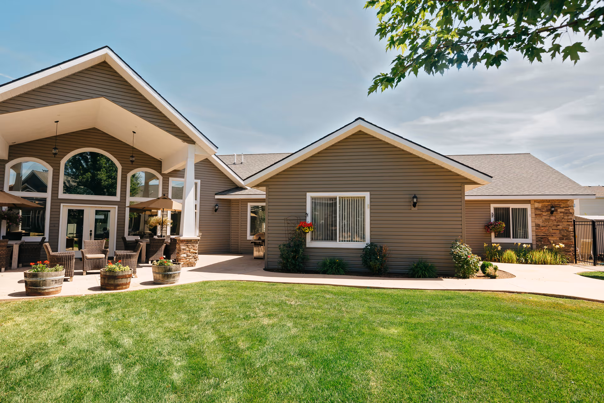 Exterior view of a single-story senior living facility building with brown siding and stone accents. The building features large windows and a covered patio area with outdoor seating and umbrellas. The foreground shows a well-maintained green lawn and some potted plants, with a clear blue sky above.