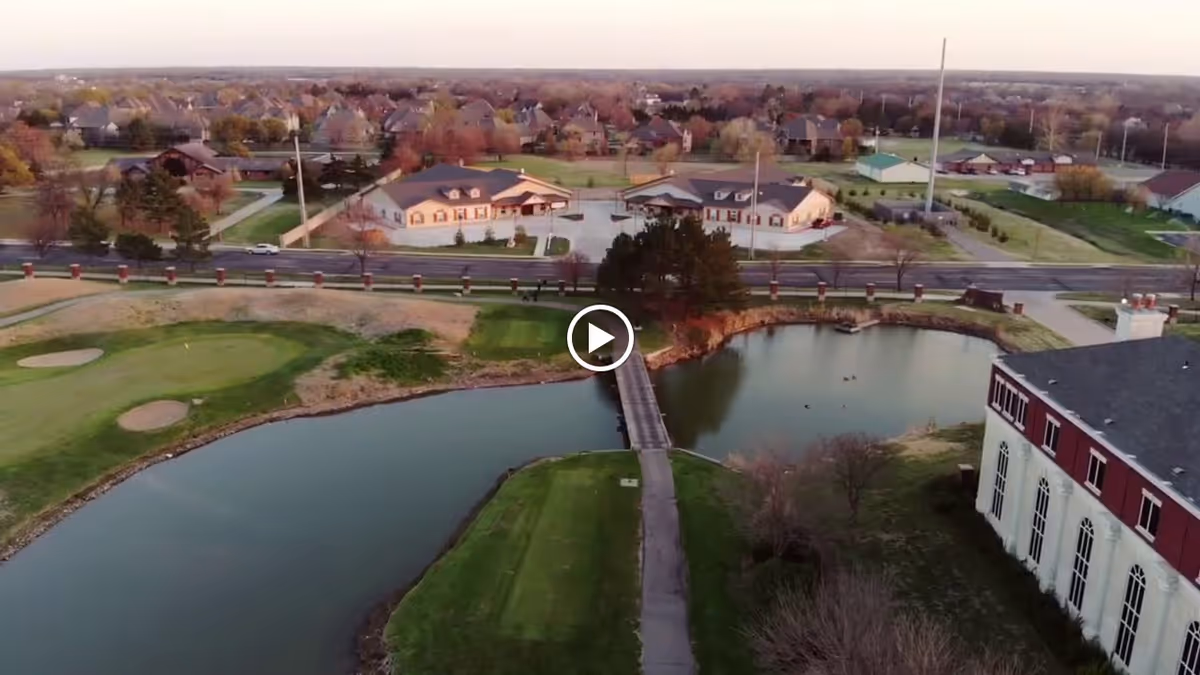 Aerial view of a suburban area featuring a small pond with a narrow bridge crossing it, surrounded by green lawns and trees. Several residential buildings and houses are visible in the background under a clear sky during sunset or early evening.
