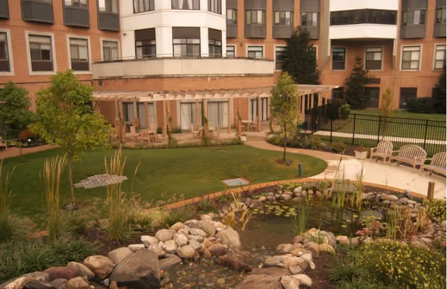 Landscaped courtyard with a small pond, pathways, benches, and surrounding multi-story brick building.
