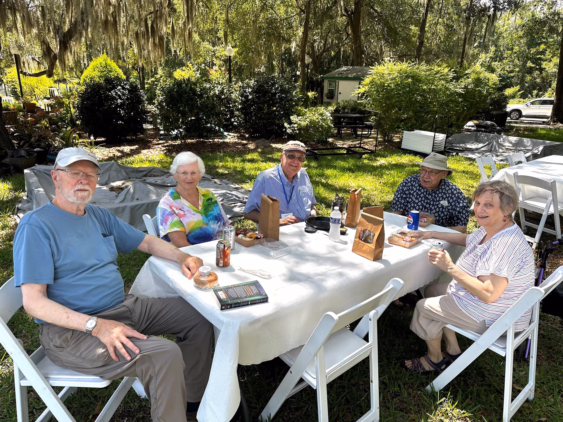 Five elderly people sitting around a white table outdoors on a sunny day, enjoying drinks and snacks. They are seated on white folding chairs with greenery and trees in the background, suggesting a garden or park setting.