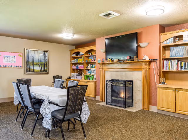 A cozy room with a rectangular table covered with a white patterned tablecloth surrounded by six chairs. The room features a fireplace with a fire burning, above which is a mounted flat-screen TV. On either side of the fireplace are wooden built-in shelves filled with books, toys, and decorative items. The walls are painted in warm tones, and there is a framed landscape painting and a bulletin board on one wall.