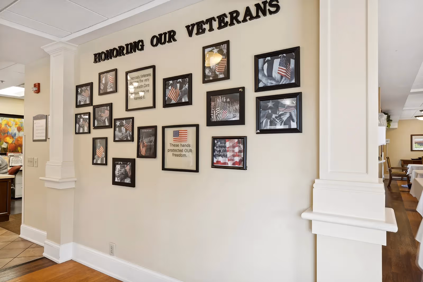 Interior wall display honoring veterans with framed photos and large 'HONORING OUR VETERANS' lettering, view into a dining area.
