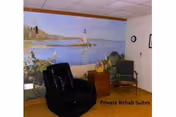 A room with a black recliner chair, a wooden side table with a plant and decorative items, and a single chair against the wall. The wall features a large mural of a coastal scene with a lighthouse and trees. The floor is wooden, and the ceiling has white tiles.