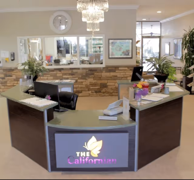 Reception desk area inside The Californian facility with a U-shaped counter, computer monitors, telephones, potted plants, a wall clock, and a chandelier hanging from the ceiling. The background includes windows and framed artwork on the walls.
