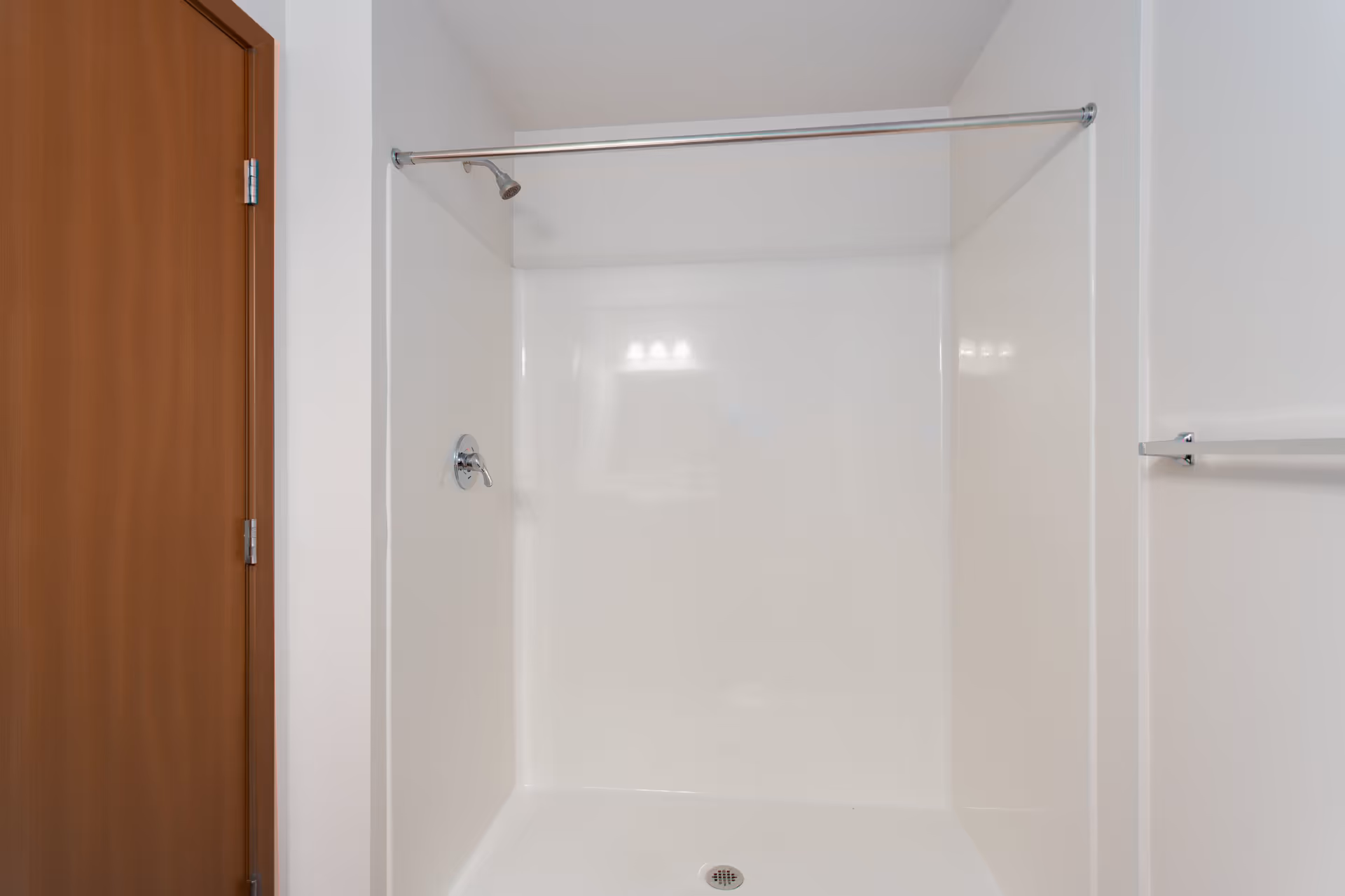 A clean, white shower stall with a chrome showerhead and a metal shower curtain rod above. To the left is a wooden door, and to the right is a chrome towel bar mounted on a white wall.