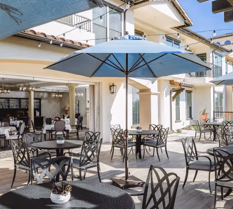 Outdoor patio with round tables, metal chairs, and blue umbrellas outside a senior living building.