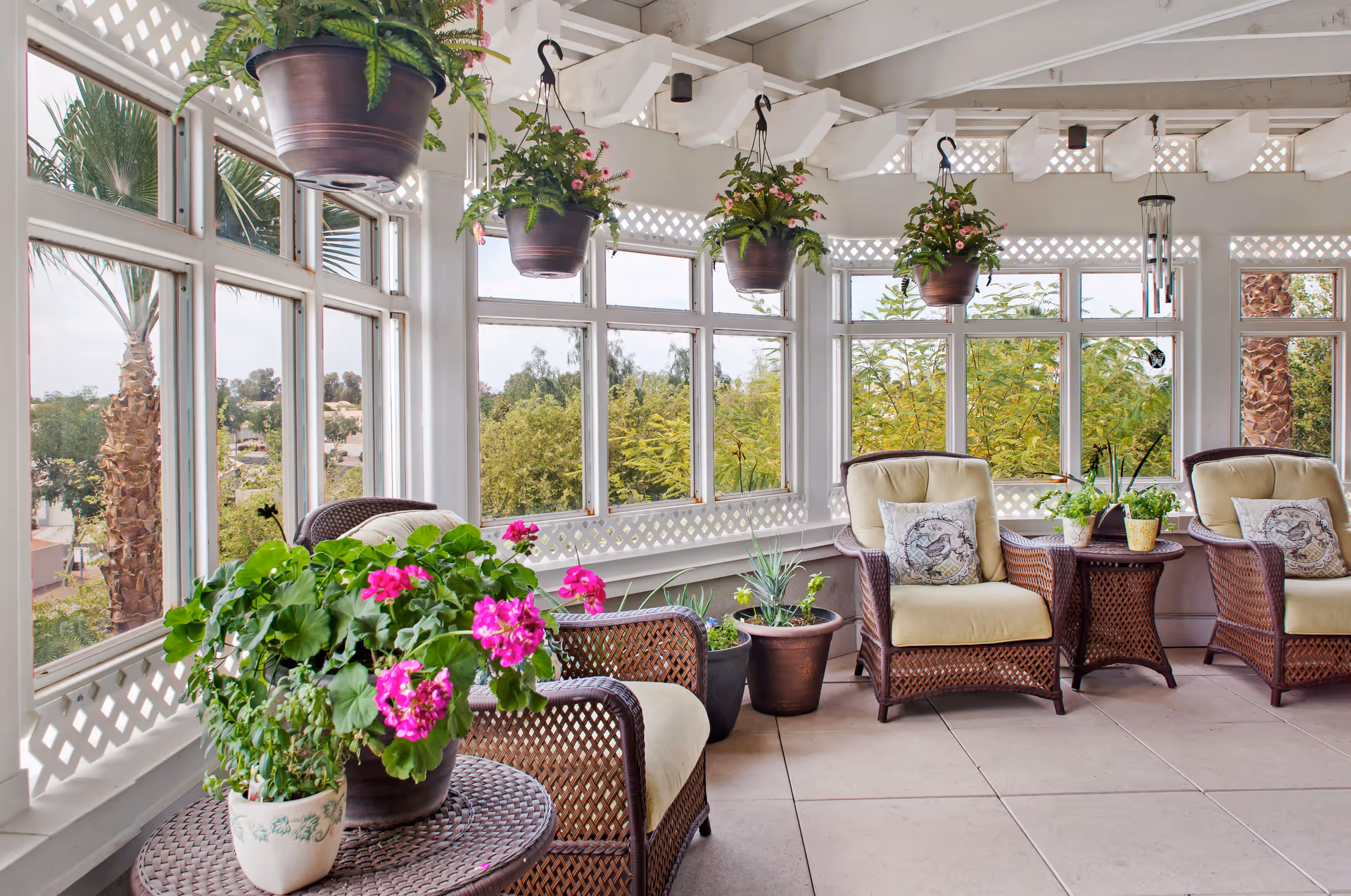 A bright and airy sunroom with large windows showing outdoor greenery. The room features wicker chairs with beige cushions and decorative pillows, a wicker side table with potted plants, and several hanging flower pots with green plants. The floor is tiled, and the ceiling has exposed white beams.
