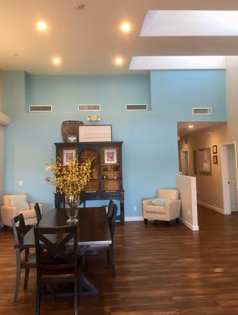 A spacious interior room with light blue walls and wooden flooring. There is a dark wooden dining table with six chairs in the center, decorated with a vase of yellow flowers. Against the wall is a dark wooden cabinet with framed pictures and decorative items. Two beige armchairs are positioned near the walls. The ceiling has recessed lighting and skylights, and a hallway is visible to the right.