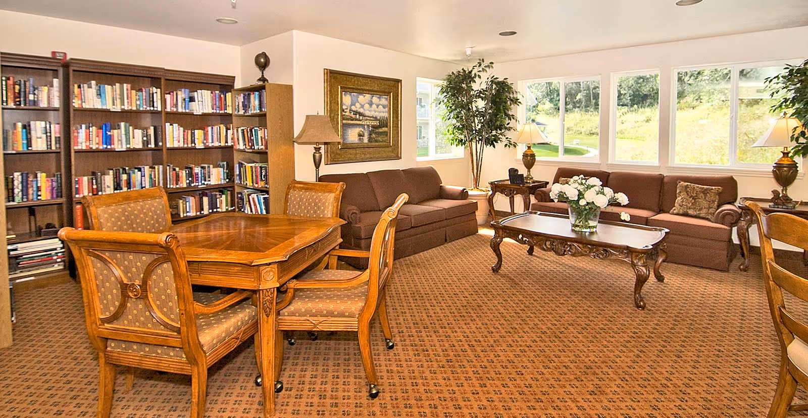 A cozy living room area with two brown sofas, a wooden coffee table with a vase of white flowers, a wooden table with four upholstered chairs, a bookshelf filled with books, two table lamps, a potted plant, and large windows showing a green outdoor view.