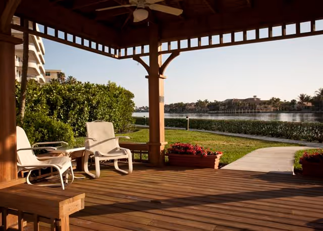 A wooden gazebo with white chairs and a small table on a wooden deck overlooking a calm body of water, surrounded by greenery and flower planters, under a clear sky.