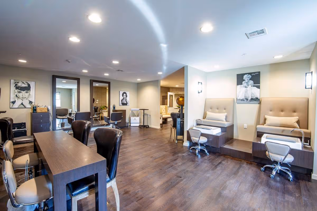 Interior view of a salon area in a senior living facility with multiple styling chairs and mirrors on the left side, and two pedicure stations with cushioned seats and foot baths on the right side. The room has wooden flooring, recessed ceiling lights, and black-and-white portraits of classic Hollywood actresses on the walls.