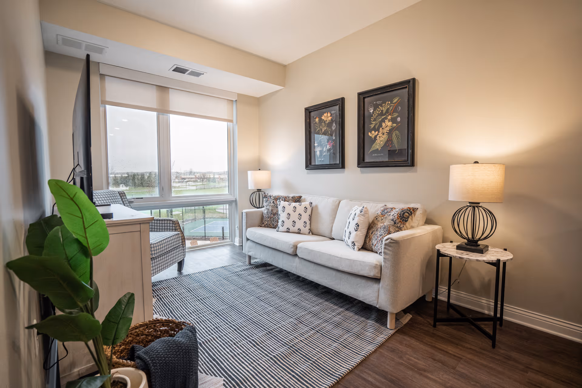 A cozy living room with a beige sofa adorned with patterned pillows, a side table with a lamp, two framed floral artworks on the wall, a large window with a view outside, a TV on a stand, a checkered armchair, a striped rug, and a potted plant in the corner.