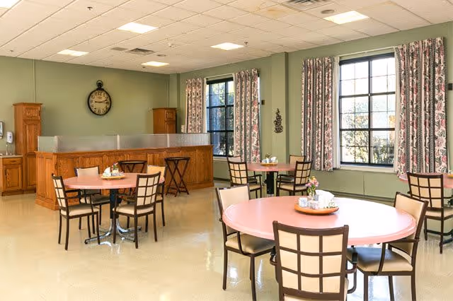 A bright dining room with round tables and chairs arranged neatly. The walls are painted light green and decorated with floral curtains on large windows letting in natural light. There is a wooden counter with a clock above it on the far wall.