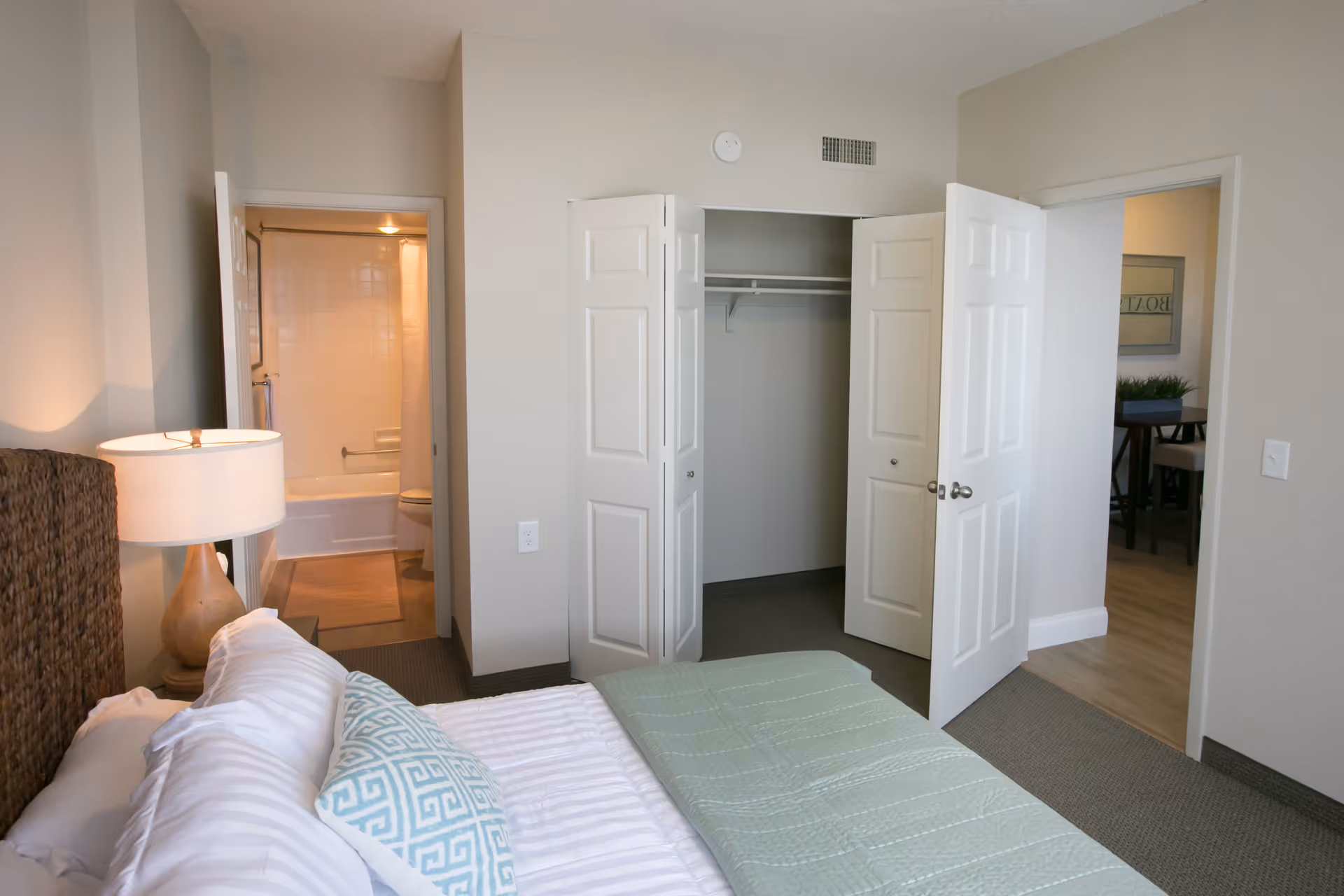 A bedroom with a neatly made bed featuring white and patterned pillows and a light green quilt. To the left of the bed is a wooden lamp on a nightstand. The room has an open closet with white double doors and an open door leading to a bathroom with a bathtub and toilet visible. Another open door leads to a dining area with a table and chairs.