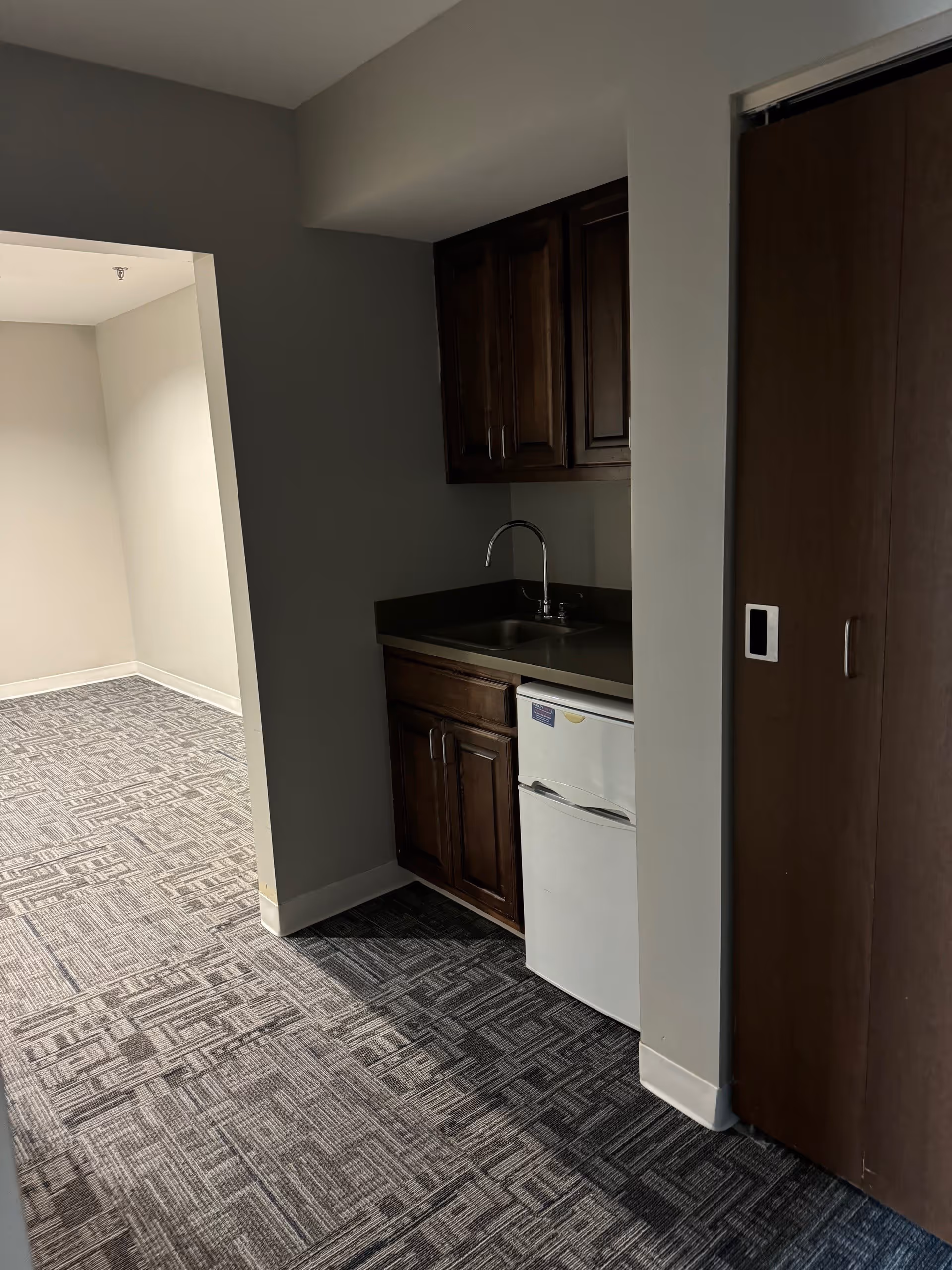 Interior view of a small kitchenette area with dark wooden cabinets, a countertop with a sink, and a compact white refrigerator underneath. The kitchenette is adjacent to a carpeted hallway with patterned gray carpet and light-colored walls.