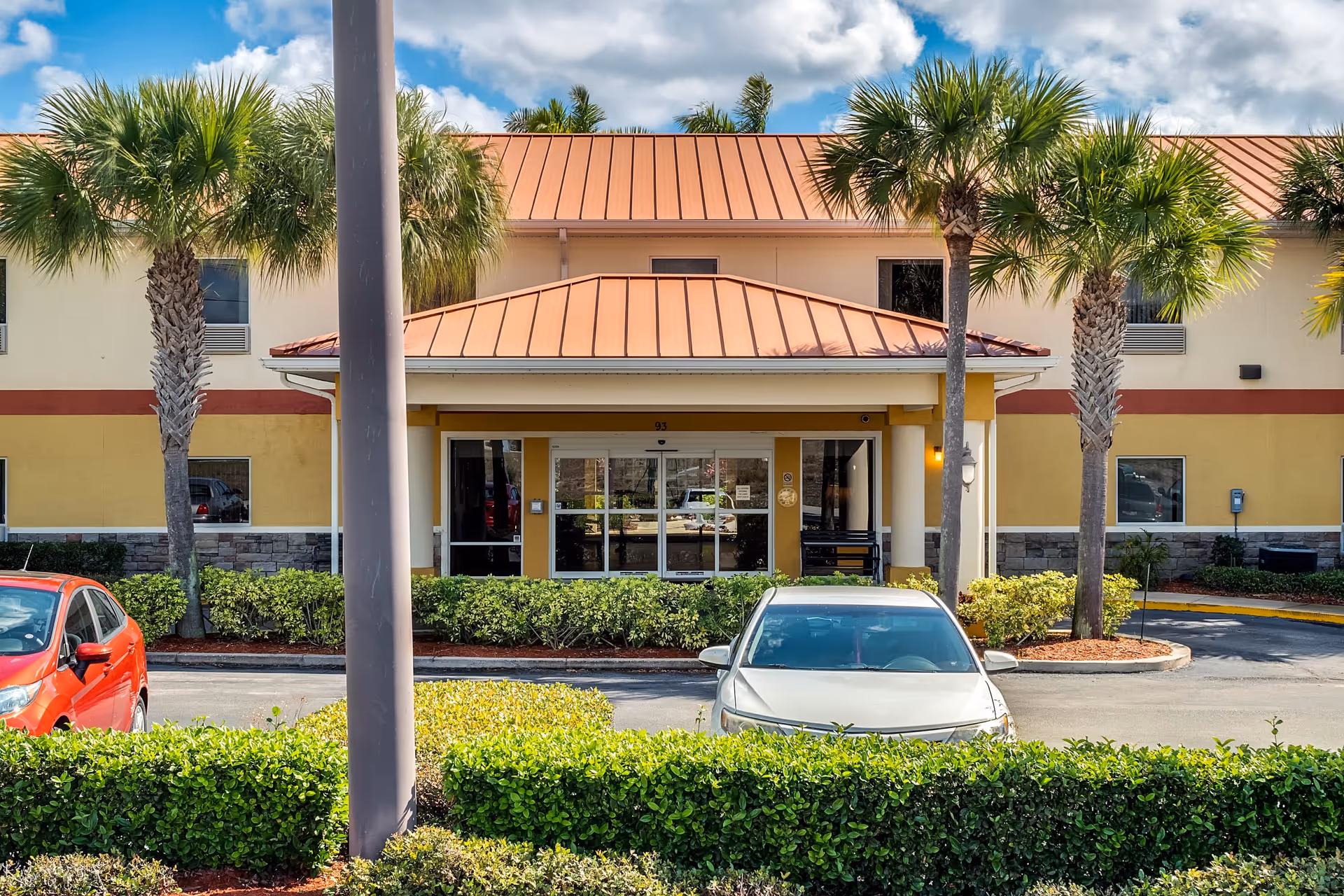 Front exterior view of Balmoral Assisted Living facility with a covered entrance, palm trees, parked cars, and a clear blue sky with some clouds.