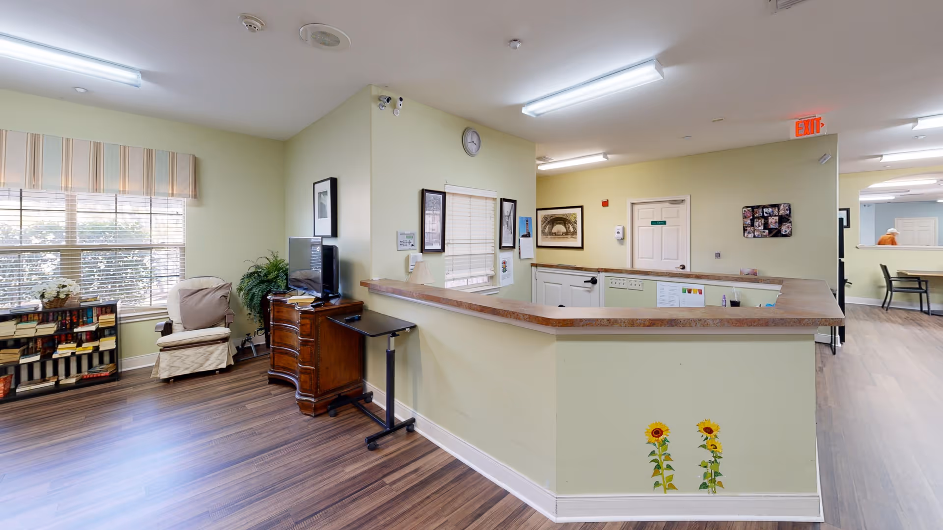 Interior view of a senior living facility reception area with a wooden floor, a light green reception desk decorated with sunflower stickers, a wooden cabinet with a TV on top, a beige armchair, a bookshelf filled with books, and windows with blinds and striped valances. The walls are light green with framed pictures and a clock above the reception window. Fluorescent ceiling lights illuminate the space.