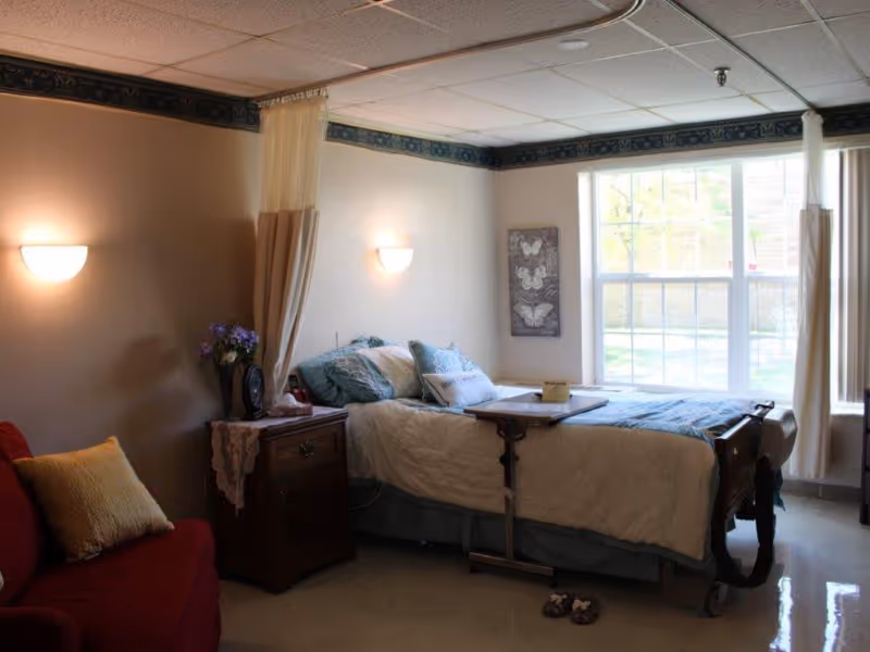 A cozy bedroom in a senior living facility featuring a hospital-style bed with blue and white bedding, a wooden bedside table with a lace doily and a vase of flowers, a red armchair with a yellow pillow, a large window letting in natural light, and a wall decoration with butterfly images.