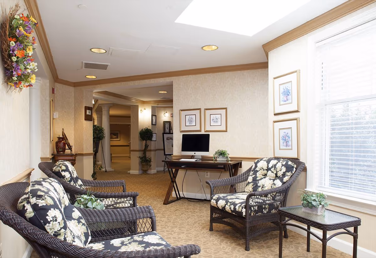 A cozy seating area in a senior living facility with three wicker chairs featuring floral cushions, a small glass-top table with a potted plant, and a desk with a computer against the wall. The room has beige walls with framed floral artwork, a large window with blinds, and a floral wreath hanging on the wall. The carpet is patterned, and the space is softly lit with recessed ceiling lights.