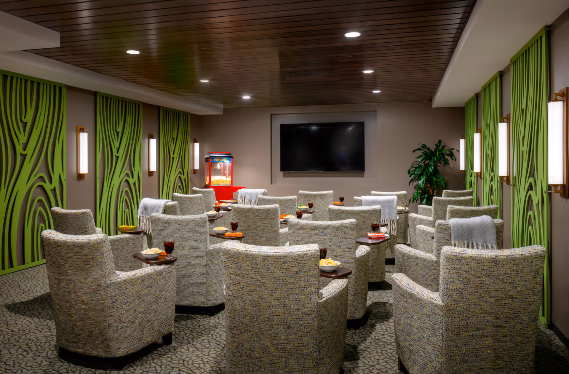 A cozy media room with multiple cushioned armchairs arranged in rows facing a large flat-screen TV mounted on the wall. Each chair has a small attached table holding snacks and drinks. The room features green decorative wall panels, modern wall sconces, a popcorn machine in the corner, and a potted plant, with a wooden ceiling and patterned carpet.