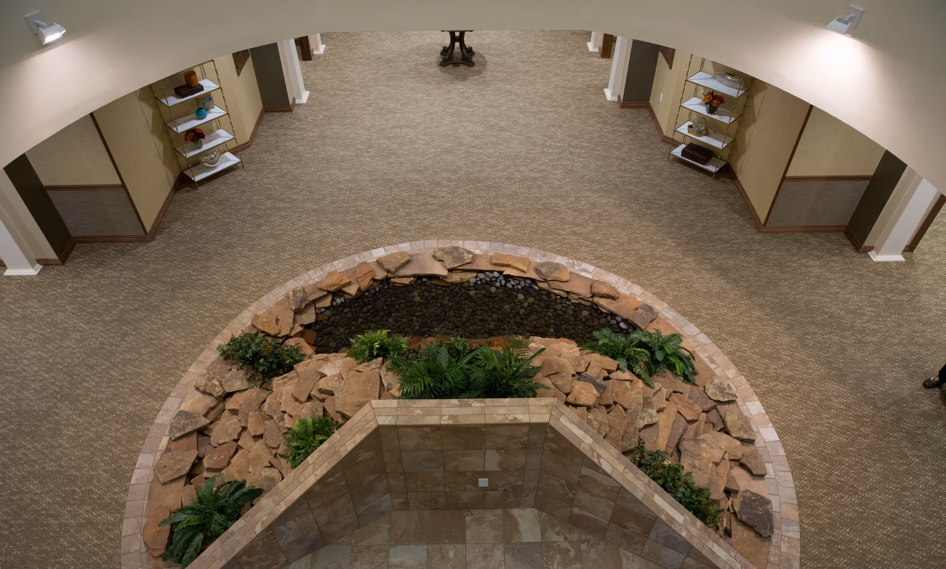 Indoor area with a circular rock and plant arrangement surrounding a small water feature, beige carpeted floor, and shelves with decorative items on the walls.