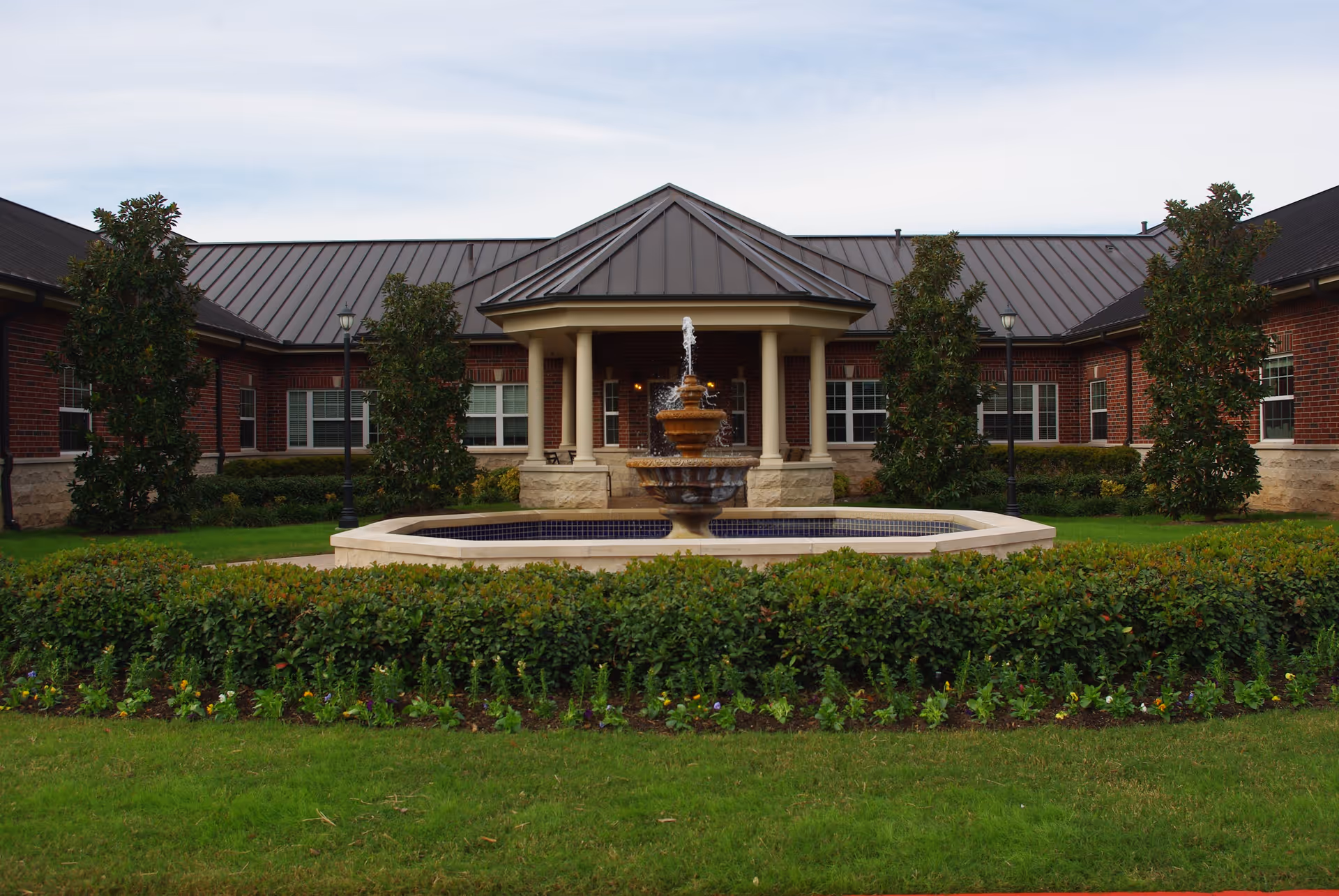 Exterior view of a single-story brick building with a metal roof, featuring a central covered entrance supported by columns. In front of the entrance is a tiered water fountain surrounded by a low hedge and green lawn, with several trees and lamp posts nearby.