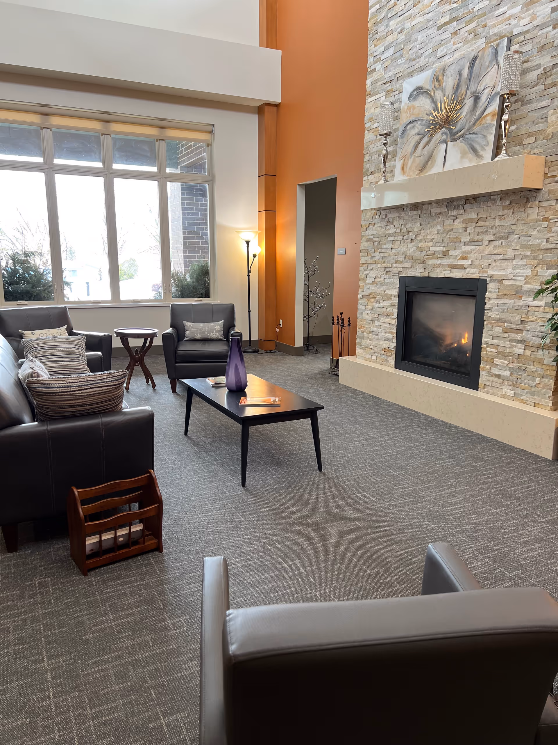 Cozy common-room lounge with leather chairs and sofas, a coffee table, large window, and a stone fireplace with artwork above.