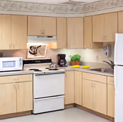 Small kitchenette with light wood cabinets, a white stove and refrigerator, microwave, coffee maker, sink, and countertop accessories.