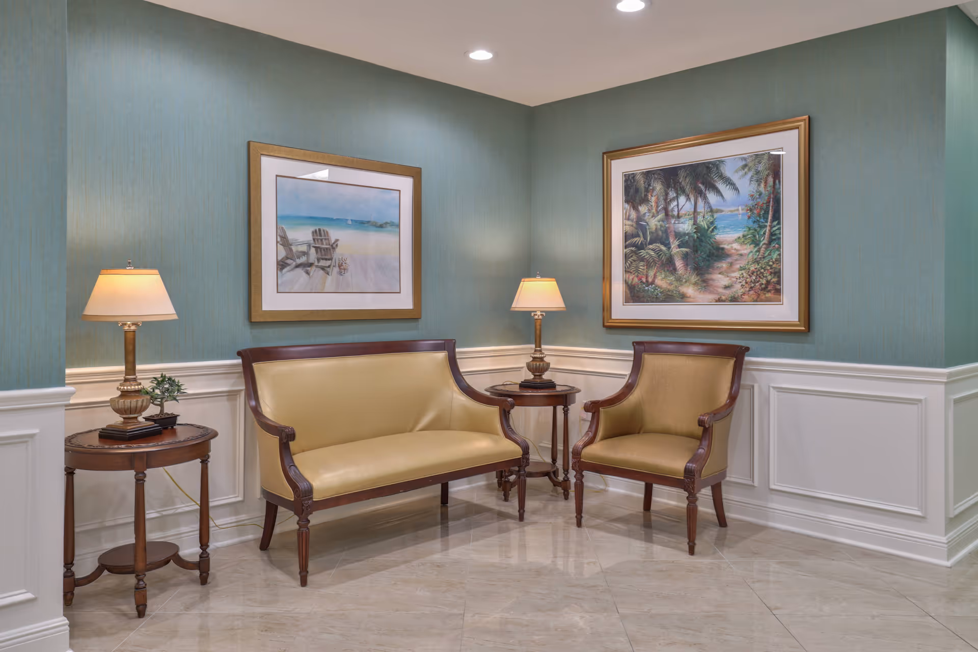 A small seating area with a beige leather loveseat and matching armchair, both with wooden frames. Two wooden side tables each hold a lamp with a beige shade. The walls are painted green with white wainscoting, and two framed paintings of beach scenes hang on the walls.