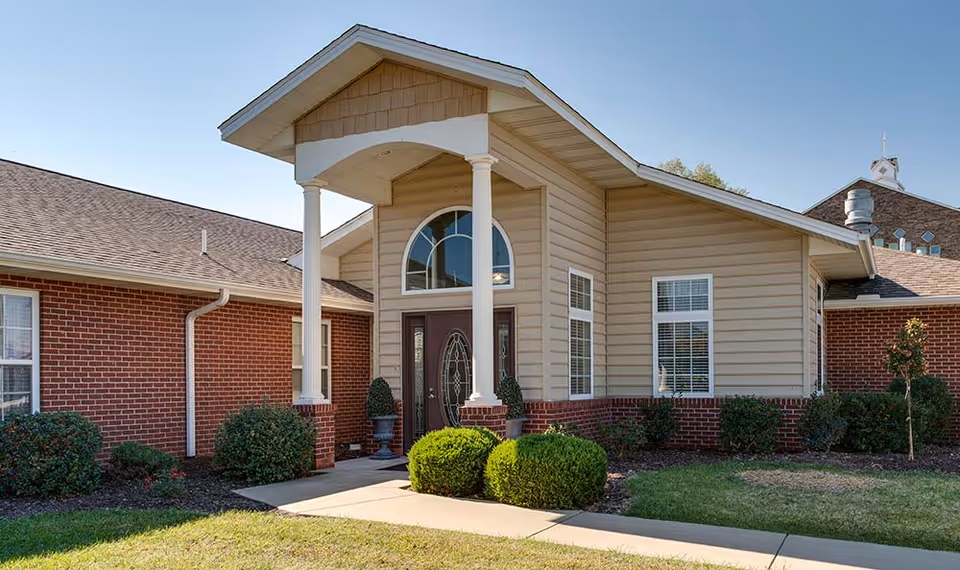 Front exterior view of a senior living facility building with a covered entrance supported by two white columns, beige siding above red brick walls, several windows, and neatly trimmed bushes and grass in the front yard.