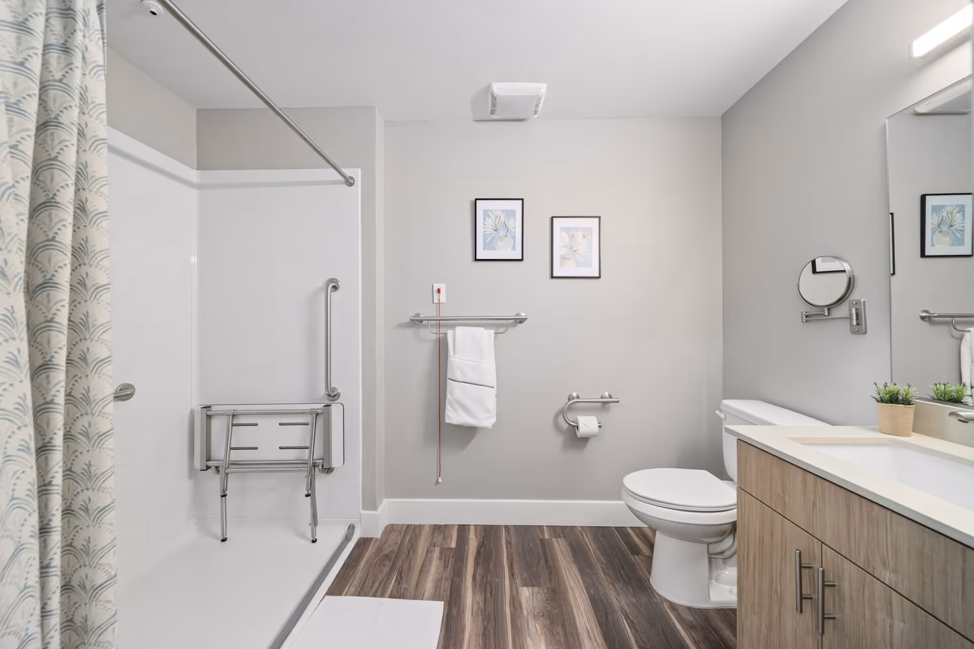 A modern bathroom with a walk-in shower featuring a foldable shower seat and grab bars. There is a white towel hanging on a rack above a red emergency pull cord. The bathroom also includes a toilet, a vanity with a sink, a small potted plant, and a round magnifying mirror mounted on the wall. Two framed floral pictures hang on the wall above the towel rack. The floor has wood-like vinyl planks.