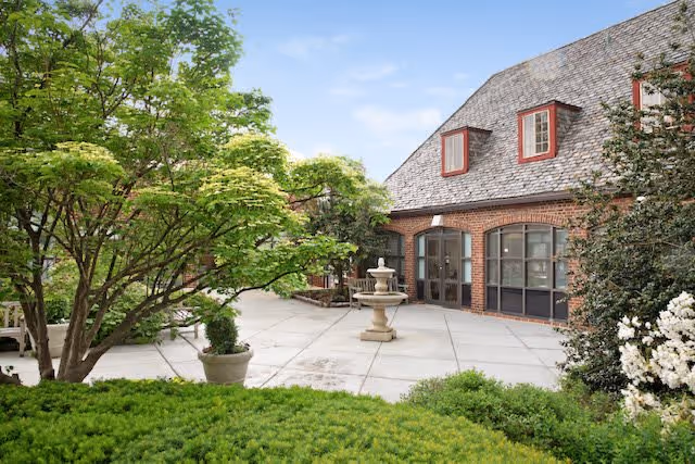 Outdoor courtyard area with a concrete patio, a central stone fountain, potted plants, and lush green trees and bushes surrounding a brick building with large windows and a shingled roof.