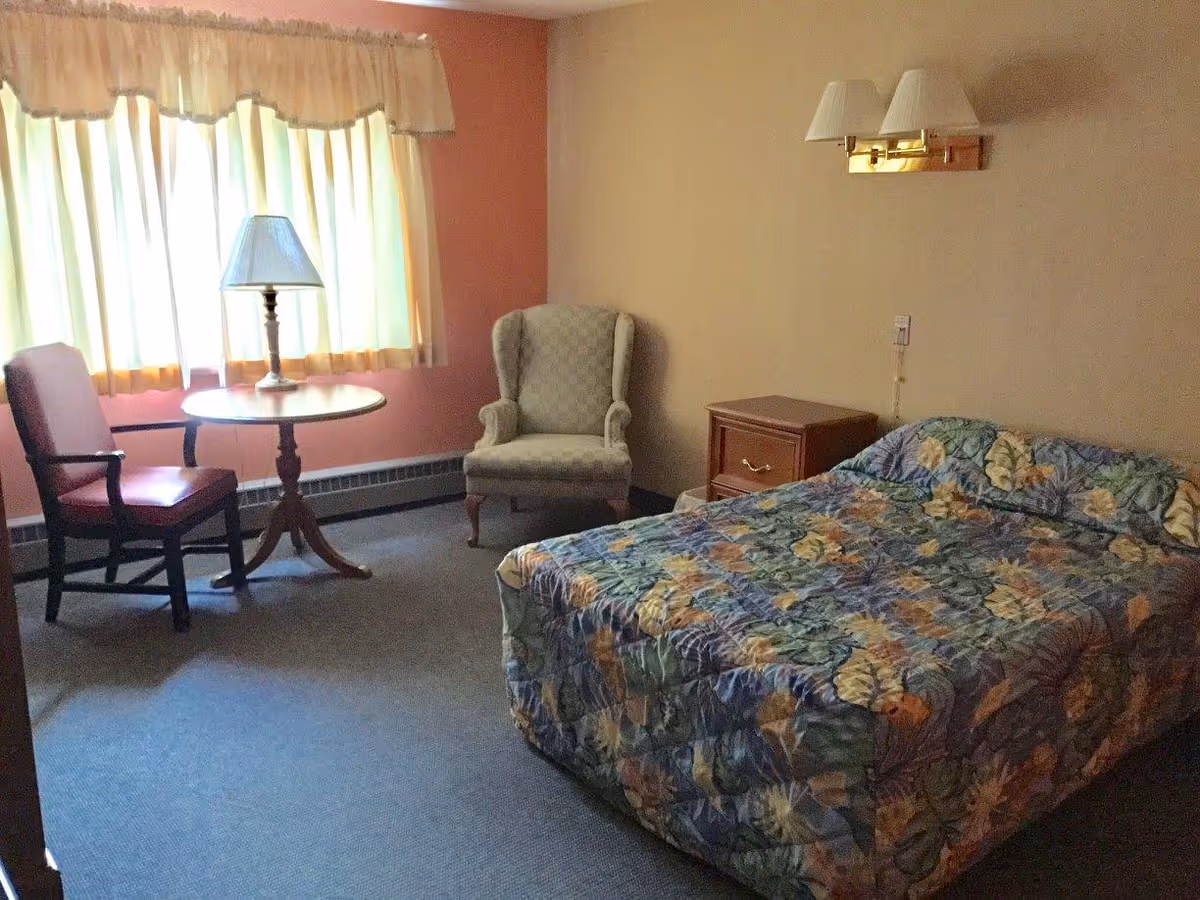 A bedroom with a bed covered in a floral patterned quilt, a wooden nightstand with a drawer, a beige upholstered armchair, a round wooden table with a lamp on it, and a wooden chair with a red cushion. The room has a window with cream-colored curtains and a wall-mounted light fixture above the bed.