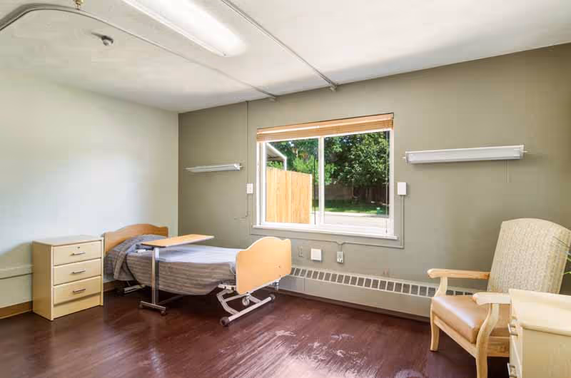 A simple, clean bedroom in a senior living facility with a single hospital-style bed, a small bedside table with drawers, a wooden armchair with a cushion, and a large window letting in natural light. The room has light green walls and dark wood flooring.