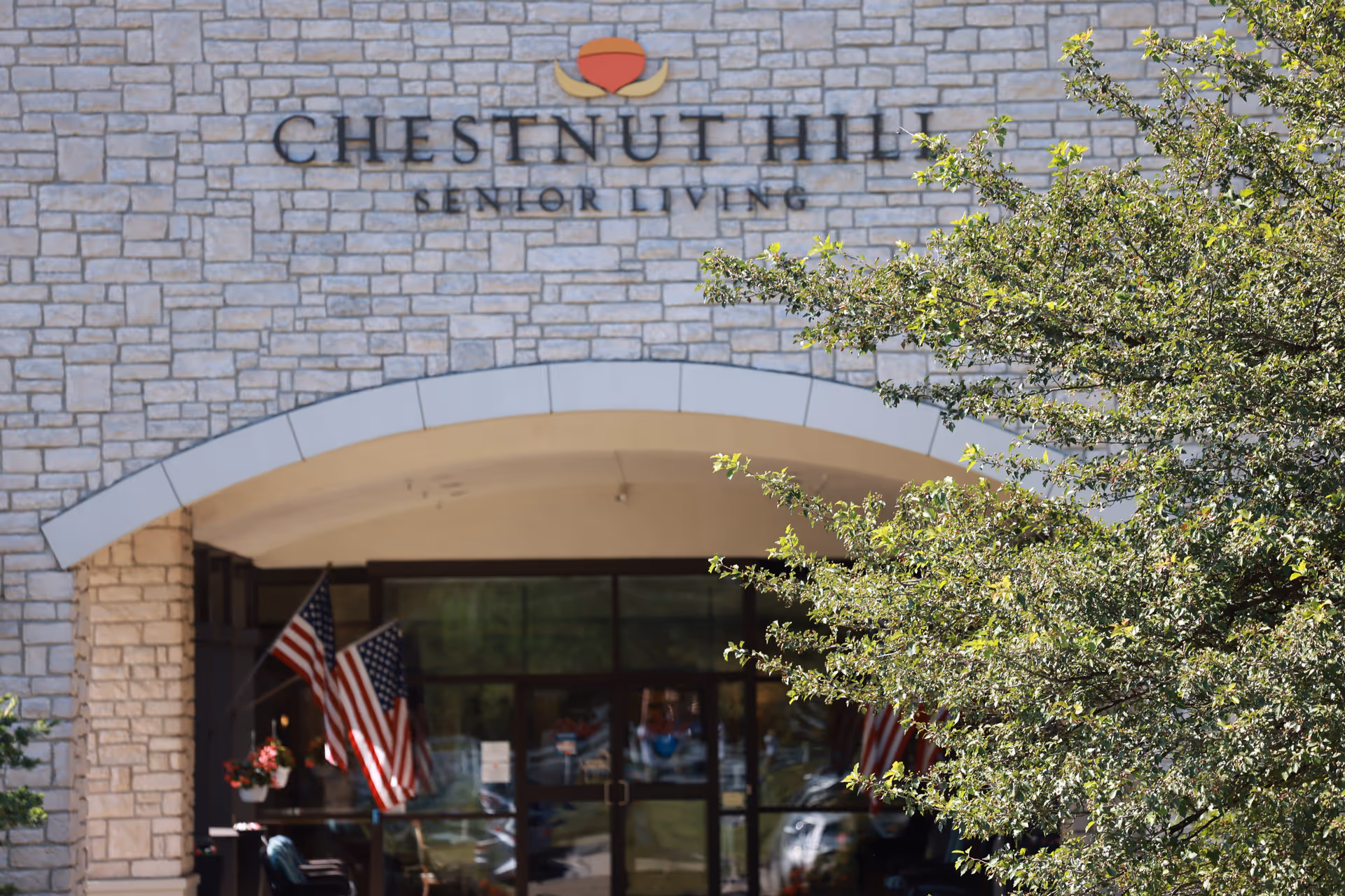 Entrance of a senior living facility named Chestnut Hill Senior Living with a stone facade, an arched doorway, two American flags displayed near the entrance, and a leafy tree partially obscuring the right side of the building.